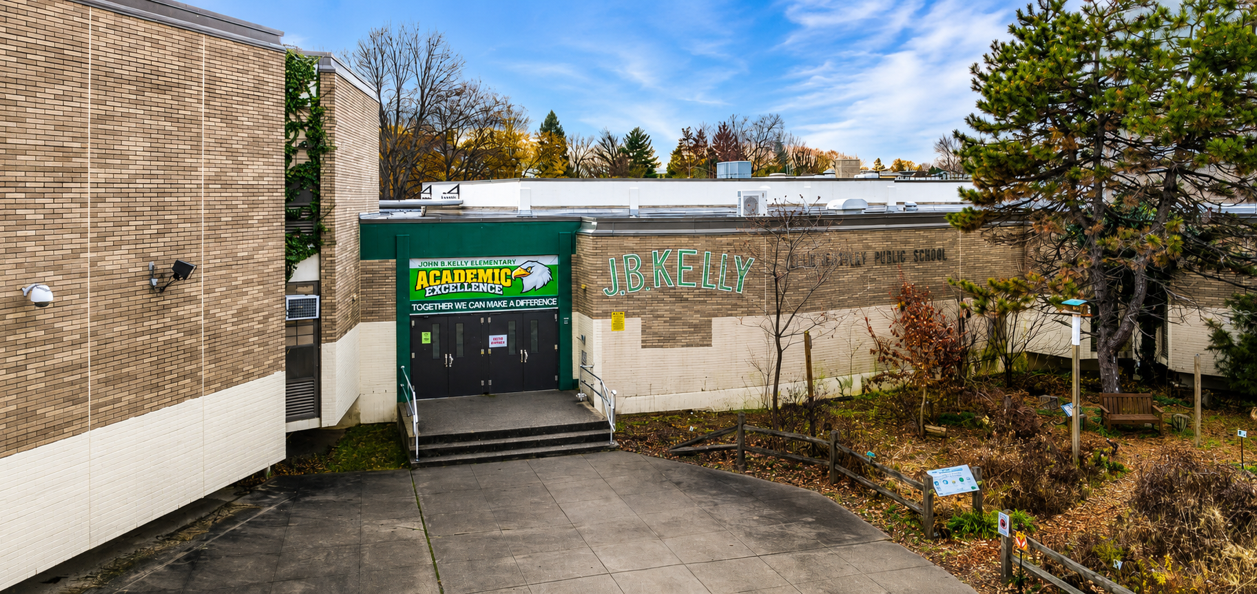 Exterior view of John B. Kelly School in Philadelphia, showing the main entrance, school signage, walkway, and surrounding trees and garden area.